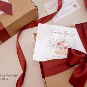 May include: Two brown gift boxes with burgundy ribbon and bows. One box has a card that says "Sending You Love" with a floral design. The text "BEAUTIFULLY PACKAGED & READY TO GIFT" is at the bottom.
