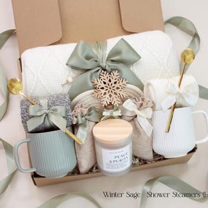 A serene holiday gift box for couples featuring a white knit blanket, two mugs, cozy socks, 2 bags of aromatherapy shower steamers, and a Peace & Purity candle, accented with a wooden snowflake ornament. The soothing sage green tones and ribbon evoke relaxation and festive calm.