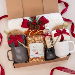 Cozy and festive Christmas gift box with gray and white mugs, caramel popcorn, festive pine and berry decor, nutcracker, red ribbons, and knit blanket — a warm and comforting holiday gift for couples.