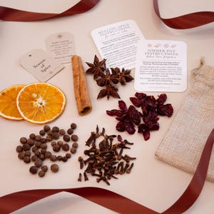 May include: A collection of mulling spices and ingredients, including dried orange slices, star anise, cinnamon sticks, peppercorns, cloves, and dried cranberries. The ingredients are arranged with instruction cards and a burlap bag, with a burgundy ribbon.