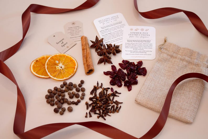 May include: A collection of mulling spices and ingredients, including dried orange slices, star anise, cinnamon sticks, peppercorns, cloves, and dried cranberries. The ingredients are arranged with instruction cards and a burlap bag, with a burgundy ribbon.