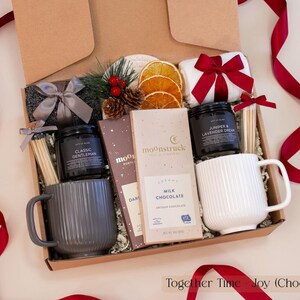 Luxurious holiday gift box for couples featuring gray and white ribbed mugs, artisan chocolate bars, pinecone and dried orange decor, cozy socks, and festive red ribbons — a warm and modern Christmas gift set.