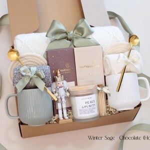 A beautifully arranged holiday gift box for couples from the Winter Sage Collection featuring a white knit blanket, two ribbed mugs in sage green and white, cozy socks tied with satin bows, Moonstruck artisan chocolate bars, a Peace & Purity candle, and a small nutcracker accent. The box is tied with a soft sage ribbon, creating a calm and elegant holiday feel.