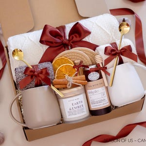 May include: A gift box filled with a white knitted blanket, two mugs, a candle, tea, honey, and cinnamon sticks. The box is decorated with burgundy bows and gold spoons. The candle reads "Earth & Ember".