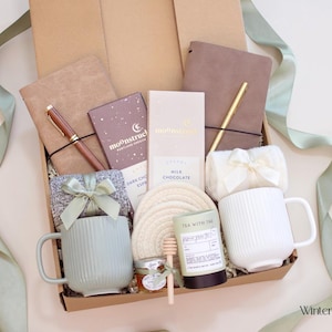 May include: A gift box filled with a variety of items, including two mugs, notebooks, chocolate bars, a pen, and tea. The mugs are light green and white, and the box is decorated with a green ribbon.