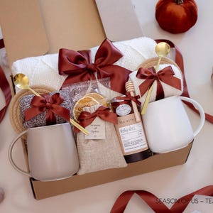 Gift box with artisan tea, pure honey, wooden dipper, dried orange slises, aromatic spice bag for simmer pots or mulling, 2 minimalist mugs, 2 long golden spoons, 2 pairs of cozy socks, soft white blanket, and 2 cotton yarn coasters, all decorated with satin ribbon in fall color, neatly arranged in a kraft gift box — a fall-inspired couples’ gift.