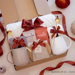 Gift box featuring rich cocoa, aromatic spice bag for simmer pots or mulling, 2 minimalist mugs, 2 long golden spoons, 2 pairs of cozy socks, soft white blanket, and 2 cotton yarn coasters, all decorated with satin ribbon in fall color, neatly arranged in a kraft gift box — a comforting couples’ gift for autumn nights.