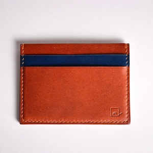 May include: A rectangular, orange leather card holder with a navy blue stripe across the top. The card holder has a stitched border and a small logo in the bottom right corner.