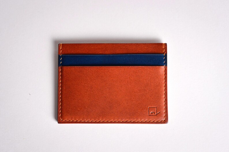 May include: A rectangular, orange leather card holder with a navy blue stripe across the top. The card holder has a stitched border and a small logo in the bottom right corner.