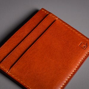 Gros plan sur un porte-cartes minimaliste en cuir brun-orangé. Le portefeuille comporte trois fentes pour cartes et une bordure cousue. Un petit logo carré est estampé sur le coin. Le fond est gris uni.