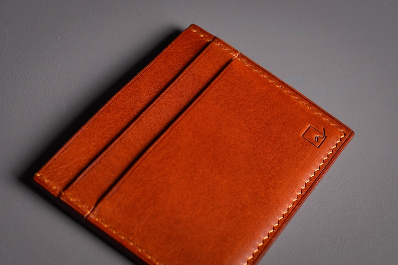 Gros plan sur un porte-cartes minimaliste en cuir brun-orangé. Le portefeuille comporte trois fentes pour cartes et une bordure cousue. Un petit logo carré est estampé sur le coin. Le fond est gris uni.