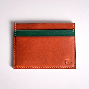 May include: A rectangular, orange leather cardholder with a green stripe. The cardholder has a stitched border and a small logo in the bottom right corner. The cardholder is approximately 4 inches wide and 3 inches tall.