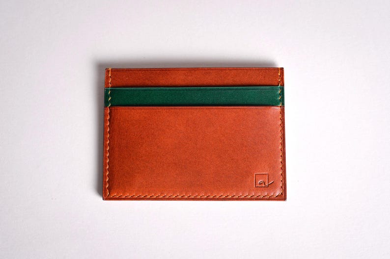 May include: A rectangular, orange leather cardholder with a green stripe. The cardholder has a stitched border and a small logo in the bottom right corner. The cardholder is approximately 4 inches wide and 3 inches tall.