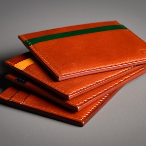 Une pile de quatre porte-cartes en cuir orange. Le porte-cartes supérieur a une bande verte, le second une bande jaune. Chaque porte-cartes est cousu avec du fil blanc. Les porte-cartes sont empilés sur une surface grise.