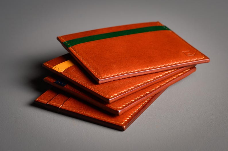 Une pile de quatre porte-cartes en cuir orange. Le porte-cartes supérieur a une bande verte, le second une bande jaune. Chaque porte-cartes est cousu avec du fil blanc. Les porte-cartes sont empilés sur une surface grise.