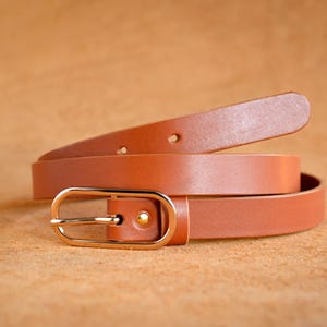 Peut inclure: Une ceinture en cuir marron clair enroulée, avec une boucle dorée. La ceinture est fine et semble être en cuir lisse. La boucle est de conception rectangulaire simple. La ceinture mesure environ 2,5 cm de large.