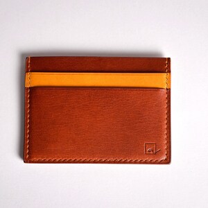 May include: A rectangular, brown leather card holder with a contrasting orange stripe. The card holder has three slots and a stitched border. A small logo is visible on the lower right corner. The product is displayed on a white background.