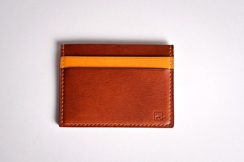 May include: A rectangular, brown leather card holder with a contrasting orange stripe. The card holder has three slots and a stitched border. A small logo is visible on the lower right corner. The product is displayed on a white background.