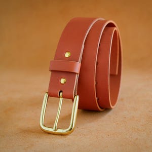 Peut inclure: Une ceinture en cuir brun-rouge enroulée, avec une boucle dorée. La ceinture a une boucle rectangulaire et deux rivets dorés près de la boucle. Le cuir semble lisse et la ceinture est enroulée pour montrer toute sa longueur.
