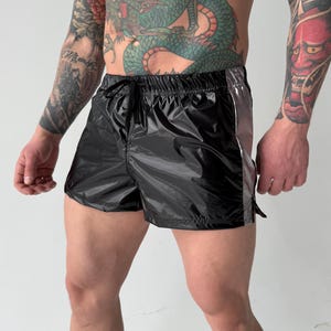 Puede incluir: Shorts deportivos negros con aspecto brillante, similar al nailon. Los shorts tienen cintura con cordón y una franja plateada en el lateral. Diseñados para actividades deportivas.