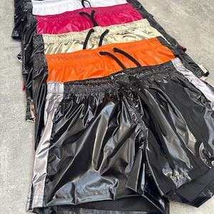 Può includere: Una pila di pantaloncini sportivi lucidi in vari colori, tra cui nero, blu navy, bianco, rosa, oro e arancione. Ogni paio ha un elastico in vita con coulisse e una striscia argentata laterale.