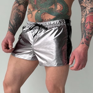 May include: Silver metallic shorts with a black stripe on the side and a drawstring waist. The shorts have a shiny, reflective appearance. The shorts are designed for swimwear or casual wear.