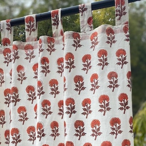 Peut inclure: Un rideau blanc orné d'un motif répétitif de fleurs orange avec des tiges et des feuilles marron. Le rideau est suspendu à une tringle noire par plusieurs pattes en tissu sur le dessus, créant une série de plis verticaux sur le tissu.