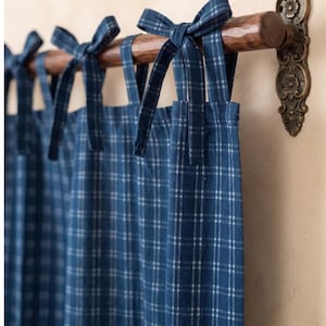 May include: A blue and white plaid curtain panel featuring fabric tie-top closures. The curtain is suspended from a dark brown wooden rod with a decorative, ornate bronze-colored metal wall bracket on the right side of the frame.
