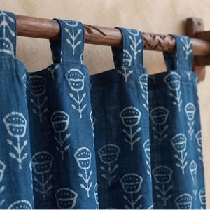 Könnte beinhalten: Ein tiefblauer Vorhang mit einem sich wiederholenden weißen Blumenmuster, der an vier Stoffschlaufen an einer dunkelbraunen Holzstange hängt. Der Stoff weist eine strukturierte Webart auf und das Blumenmotiv besteht aus stilisierten Blüten.