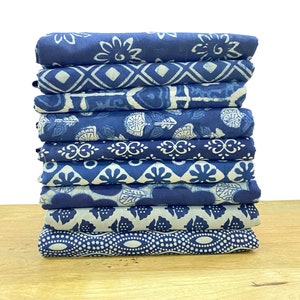 Op de afbeelding: Een stapel van tien blauwe en witte katoenen vierkanten met verschillende patronen. De stof is gevouwen en gestapeld op een houten oppervlak.