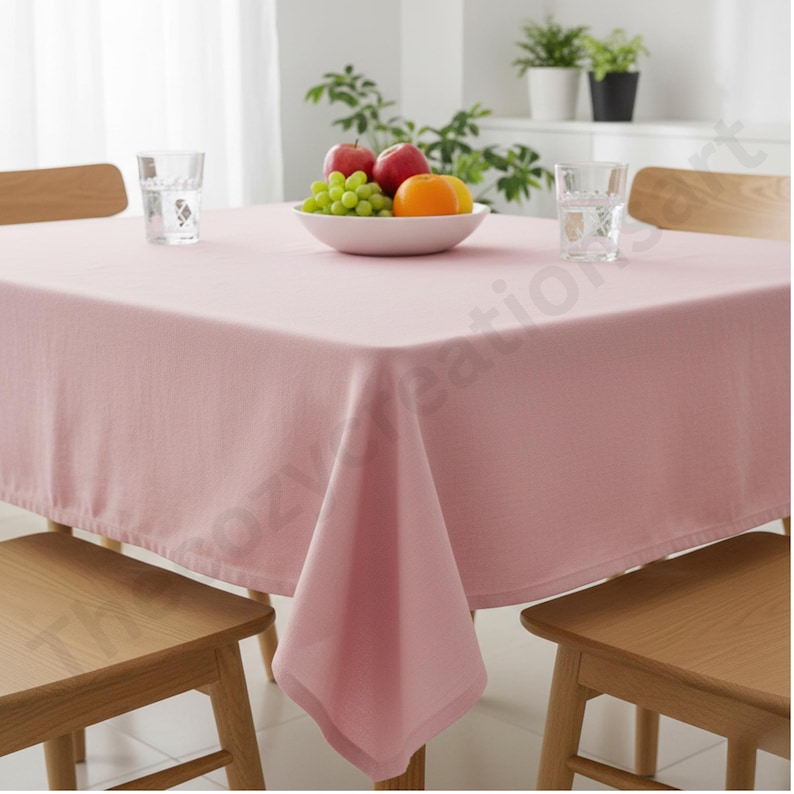Puede incluir: Un mantel cuadrado rosa claro cubre una mesa de madera, con dos sillas de madera visibles. Un cuenco de fruta, que incluye manzanas, uvas y una naranja, se encuentra en el centro. Dos vasos de agua tambi&eacute;n est&aacute;n sobre la mesa.
