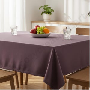 Puede incluir: Un mantel rectangular de color ciruela cubre una mesa de madera. Un cuenco blanco con fruta, incluyendo manzanas, uvas y una naranja, est&aacute; en el centro. Dos vasos de agua estampados est&aacute;n sobre la mesa. Sillas de madera a los lados.