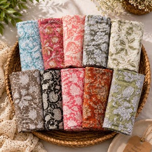 May include: A collection of ten rectangular cotton napkins arranged in a round woven basket. Each napkin features a distinct floral block print pattern in various colors including blue, pink, red, brown, black, and green with white floral details.