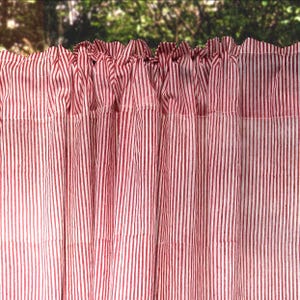 May include: Red and white striped curtain panel. The fabric is gathered at the top, creating a ruffled effect. Vertical stripes run the length of the panel. The curtain is made of a lightweight fabric, suitable for a window treatment. The background is blurred.