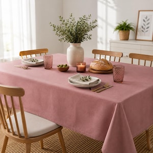 May include: A solid dusty rose tablecloth draped over a rectangular dining table. The table is set with a white ceramic vase containing green foliage, a round loaf of bread on a wooden board, two textured mauve glasses, and folded napkins.