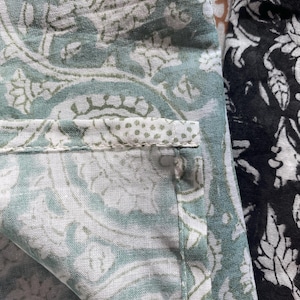 May include: A textile fabric featuring a sage green and white paisley pattern with a visible stitched hem. Adjacent to this is a section of black fabric with a white floral print. The material appears to be a lightweight woven cotton with visible seams.