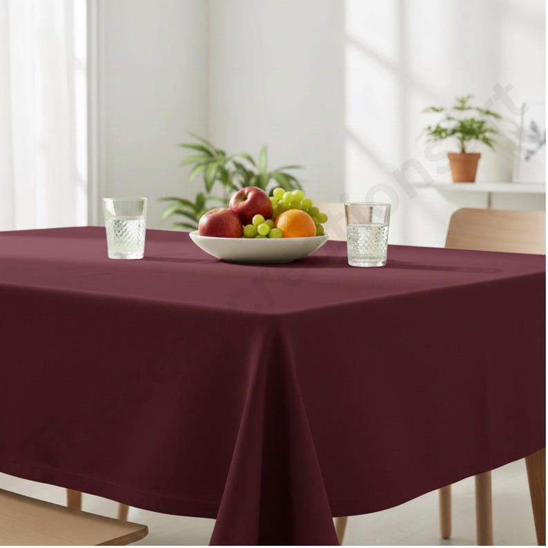Puede incluir: Un mantel cuadrado de color burdeos cubre una mesa de madera. Un cuenco blanco con fruta, incluyendo manzanas, uvas y una naranja, est&aacute; en el centro. Dos vasos de agua transparentes est&aacute;n a ambos lados del cuenco. Una planta en maceta est&aacute; en el fondo.