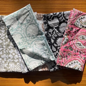 May include: Four folded fabric napkins featuring distinct patterns. One has a beige geometric print, another a light green paisley design, a third a black and white floral motif, and the fourth a pink paisley pattern with green and white accents.