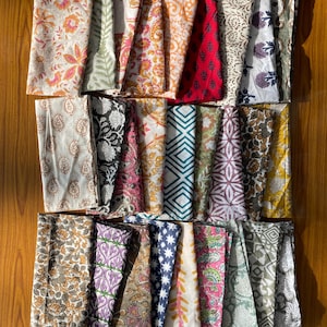 May include: A collection of rectangular fabric napkins featuring a variety of block-printed patterns in diverse colors including pink, orange, red, blue, green, yellow, and purple, arranged in three horizontal rows on a wooden surface.