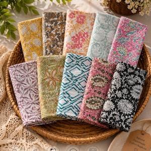 May include: A collection of ten folded cotton napkins featuring diverse floral, geometric, and paisley patterns in yellow, gray, pink, blue, green, and black. A brown tag reads: Natural Handmade Cotton Napkins, Reusable, Eco-Friendly, Sustainable.