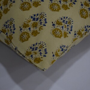 Pode incluir: Close-up de uma almofada de cor creme com um padr&atilde;o floral azul e amarelo. A almofada &eacute; feita de um tecido macio e texturizado.