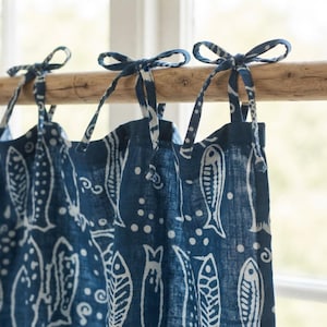 Pode incluir: Uma cortina azul-escura com um padrão de peixes brancos e pequenos pontos. A cortina está suspensa numa vara de madeira castanho-clara por três pares de fitas de tecido atadas em laços no topo, criando um aspeto franzido e suspenso.