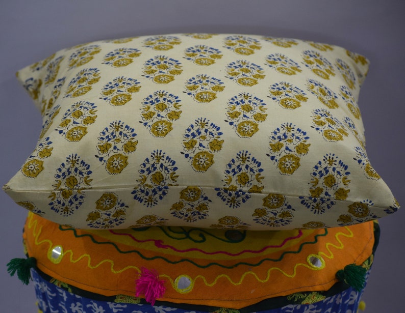 Pode incluir: Uma almofada de cor creme com um padr&atilde;o floral em tons de amarelo e azul. A almofada est&aacute; colocada em cima de um coj&iacute;n colorido com padr&atilde;o.