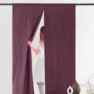 May include: Plum-colored linen curtains hanging from a dark rod. One curtain is partially drawn back, revealing a person in a white outfit. A decorative vase and plant are visible in the lower right corner. The curtains have a textured appearance.