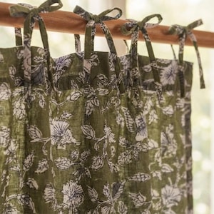 May include: A curtain panel featuring an olive green base colour with a white floral pattern. The curtain is suspended from a wooden rod by several fabric ties that are knotted at the top, creating a gathered look across the upper edge of the fabric.