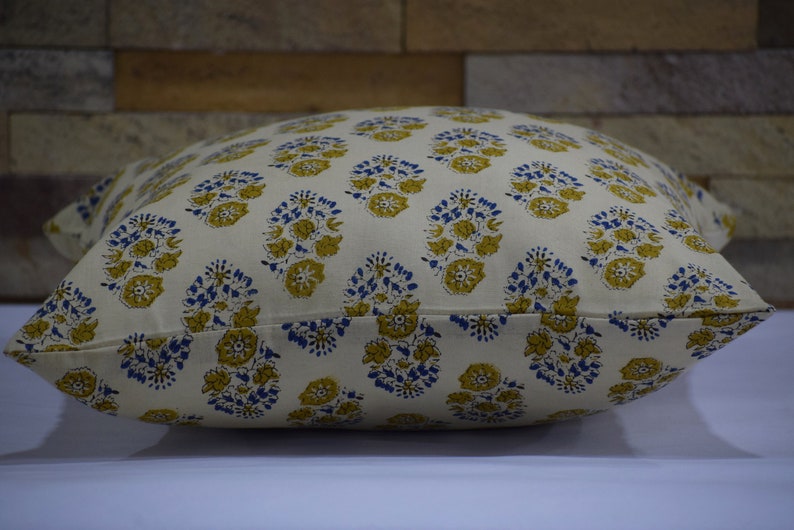 Pode incluir: Uma capa de almofada branca com um padr&atilde;o floral em tons de azul e amarelo. O padr&atilde;o apresenta pequenos motivos repetidos de flores e folhas.