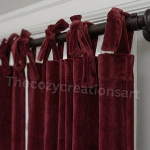 May include: Burgundy velvet curtains with tie-top closures. The curtains are hanging from a dark brown rod. The text "The cozy creations art" is visible on the lower portion of the curtains.