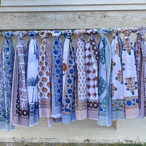 K&ouml;nnte beinhalten: Eine Sammlung von 10 wei&szlig;en Baumwollschals mit blauen und braunen Blockdruckmustern. Die Schals h&auml;ngen an einer Holzstange vor einer wei&szlig;en Backsteinwand.