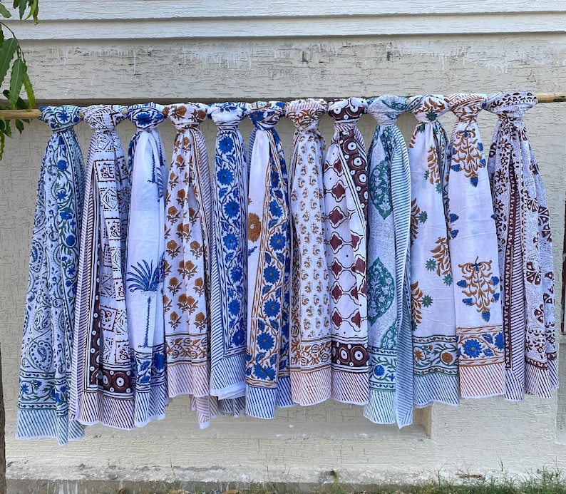 K&ouml;nnte beinhalten: Eine Sammlung von 10 wei&szlig;en Baumwollschals mit blauen und braunen Blockdruckmustern. Die Schals h&auml;ngen an einer Holzstange vor einer wei&szlig;en Backsteinwand.