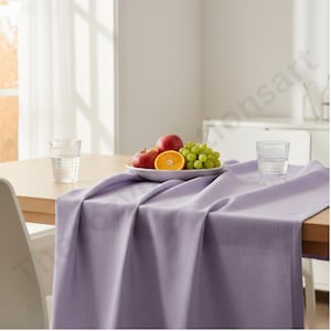 Puede incluir: Un camino de mesa lila sobre una mesa de madera. Un plato blanco con manzanas, uvas y media naranja. Dos vasos transparentes sobre la mesa. La habitaci&oacute;n es luminosa.